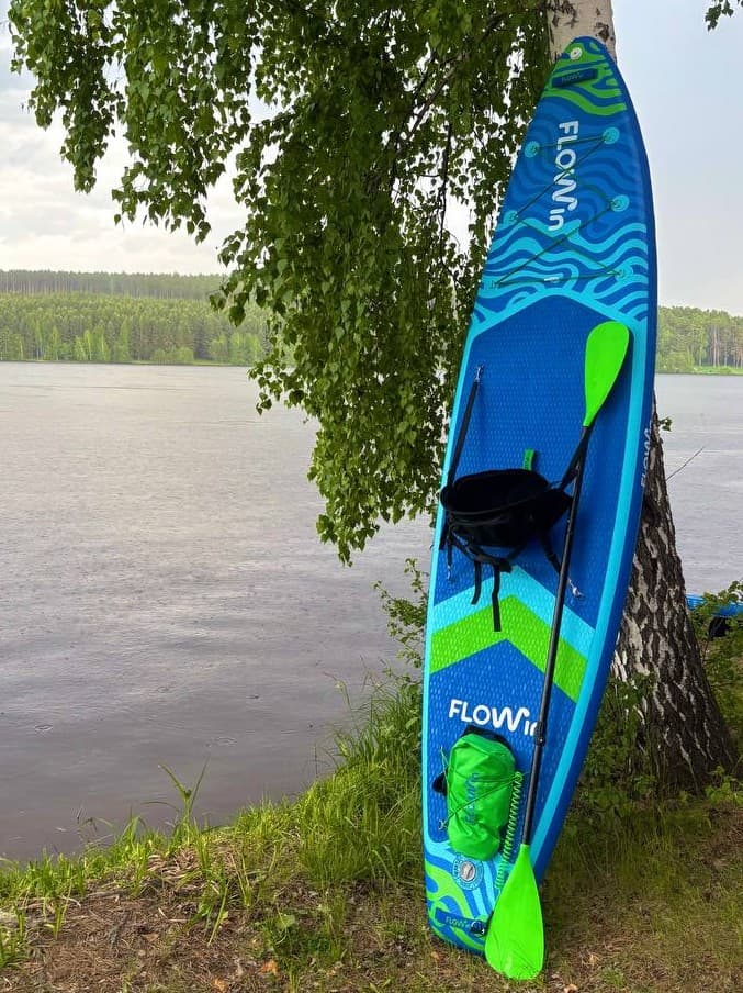 Крепление для весла на сапборд: надувная доска Flowin стоит у берёзы на берегу реки, на палубе пристёгнуто сиденье, у хвоста под эластичным шнуром лежит зелёный гермомешок, рядом с доской стоит весло с зелёной лопастью